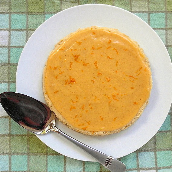 Overhead view of a whole pumpkin butter Japanese cheesecake on a white platter. Silver cake server on side of platter. Tablecloth has squares of different shades of pastel greens and white with grey dividers. 