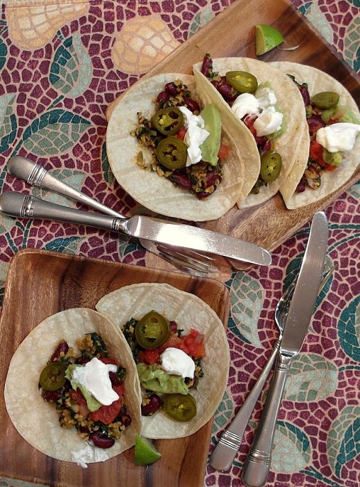 Veggie oat taco mince tacos. Three on one plate, two on another. Plated on wooden square plates. Served on a table with mosaic design table cloth and silver forks and knives.