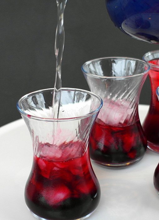Hibiscus rose sharbat pouring into Turkish clear glasses. Beverage is deep red over ice. 