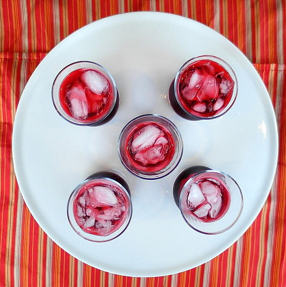 Hibiscus rose sharbat over ice in glasses. Picture is taken from above showing the red beverage. 5 glasses are on a white platter which is on a tablecloth of red yellow and white stripes.