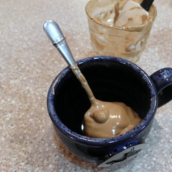 A blue earthenware mug with a thick foam of instant coffee, sugar and water in the bottom with a metal spoon.