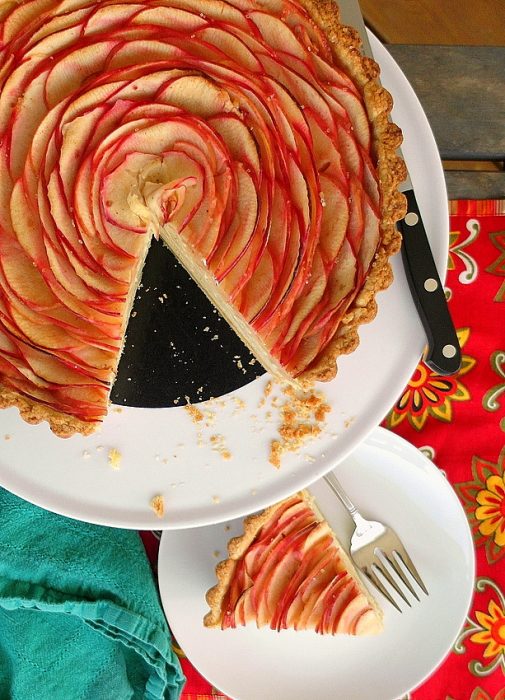 Overhead shot of chai apple cheesecake tart with concentric rings of thin apple slices and a cut slice of the tart on a separate plate.