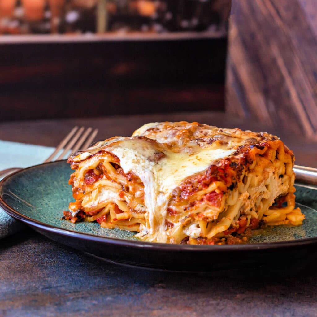 A square slice of lasagna on a plate with some melted cheese running down one side of the slice.