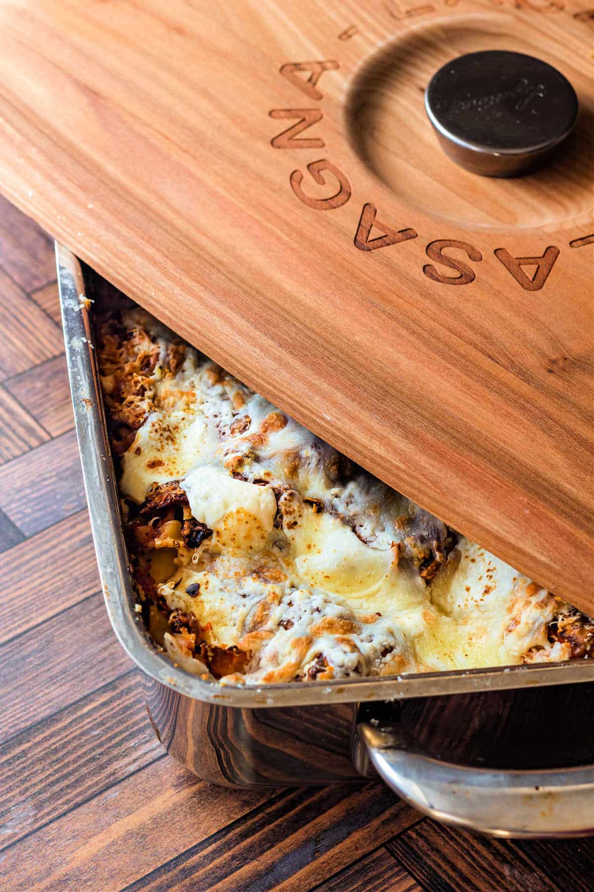 An overhead shot of a metal pan of sausage lasagna with cheese on top. There is a wooden lid for the pan, and the word "lasagna" is wood-burned into the wood around the knob. It is set askew on the pan so you can see a large corner of the lasagna peeking out.