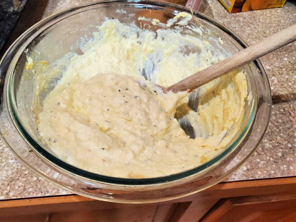 A glass bowl of ricotta cheese mixed with egg, other cheeses, and seasonings with a wooden spoon in it.