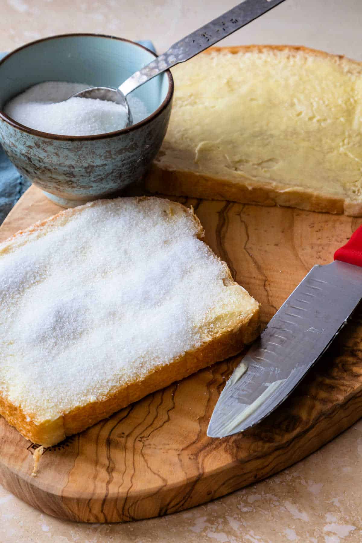 Aslice of bread completely covered with a layer of granulated sugar, another slice slathered in butter, a spreading knife, and a small blue bowl with sugar in it.