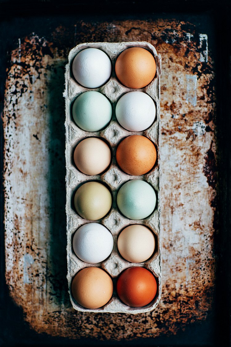 An overhead shot of a dozen different colored eggs in a carton on a distressed background.