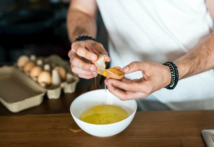A chef using egg shells to separate whites from yolks into a white bowl. A carton of brown eggs sits in the background.