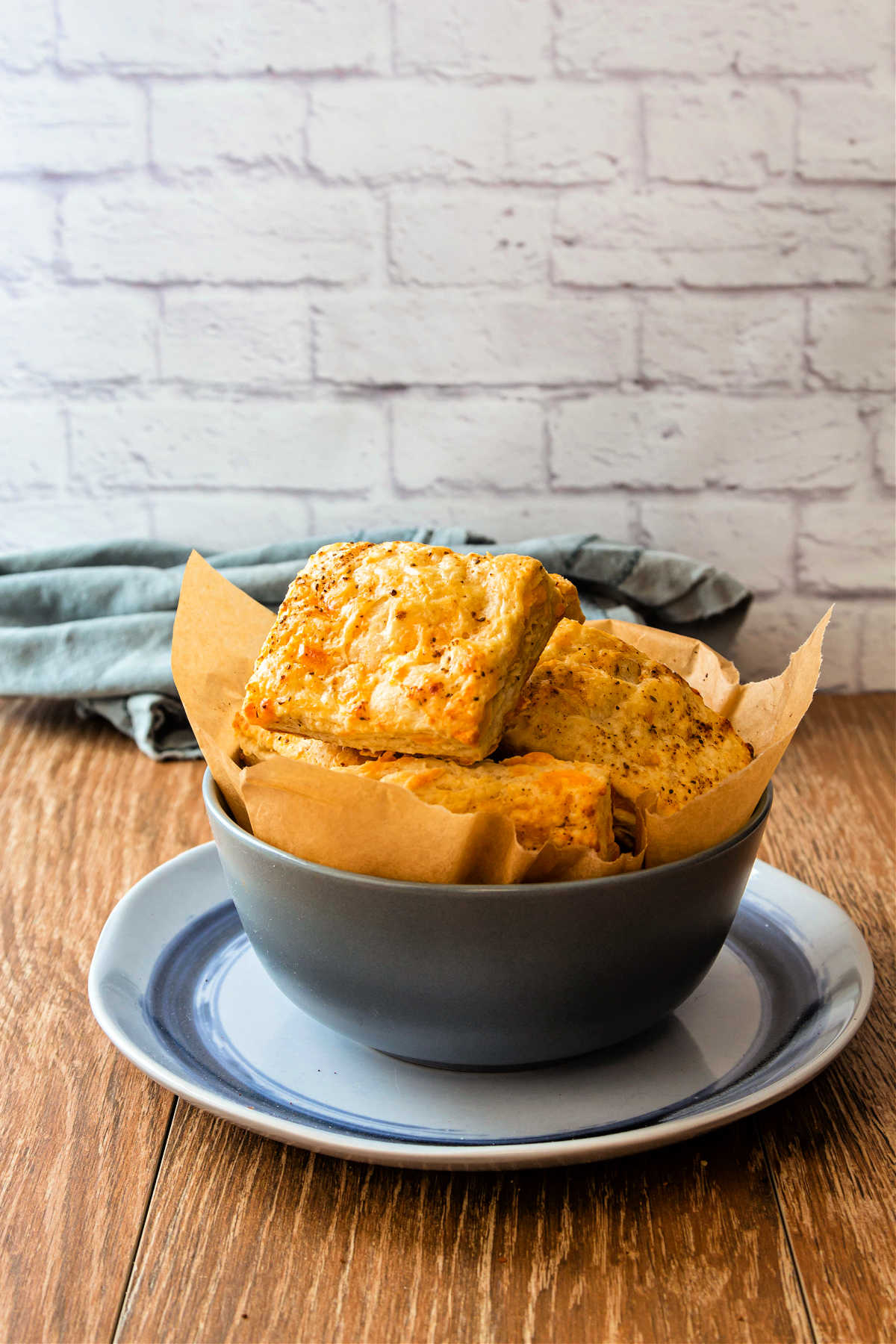 A slate-colored bowl with square cheddar biscuits in it. The bowl is on a blue plate and shot against a backdrop of white bricks.