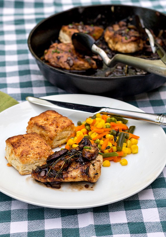 Chicken thigh with mushrooms and thyme, 2 biscuits, and mixed vegetables on a white plate with a cast iron skillet in the background with more thighs.
