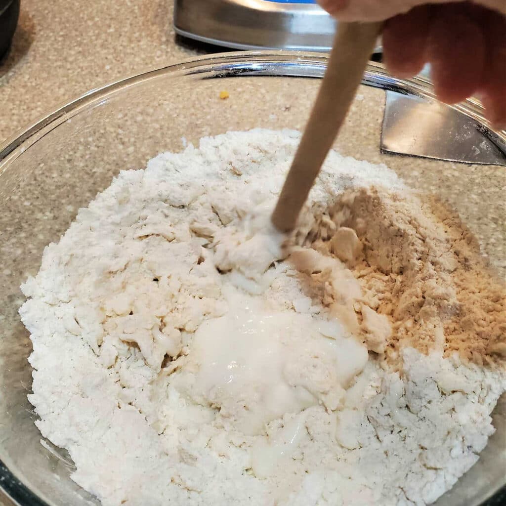A close up shot of a glass bowl of flour/butter mixture and stirring in buttermilk with the handle of a wooden spoon.