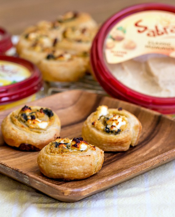 3 puff pastry hummus pinwheels on a wooden plate with containers of Sabra hummus in the background.