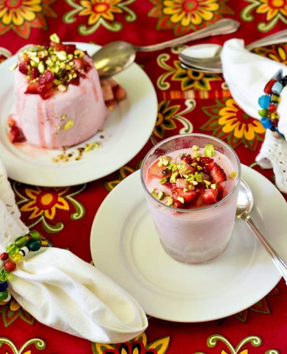 A clear glass of strawberry kulfi topped with diced strawberry and chopped pistachio served on a white plate with a silver spoon.