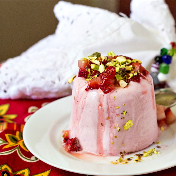 An unmolded dome shaped kulfi on a plate with strawberries and pistachio as garnish.