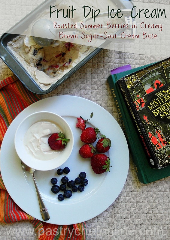 A metal loaf pan with fruit dip ice cream  being scooped out. A white plate with the ice cream, berries and some books to the side. 