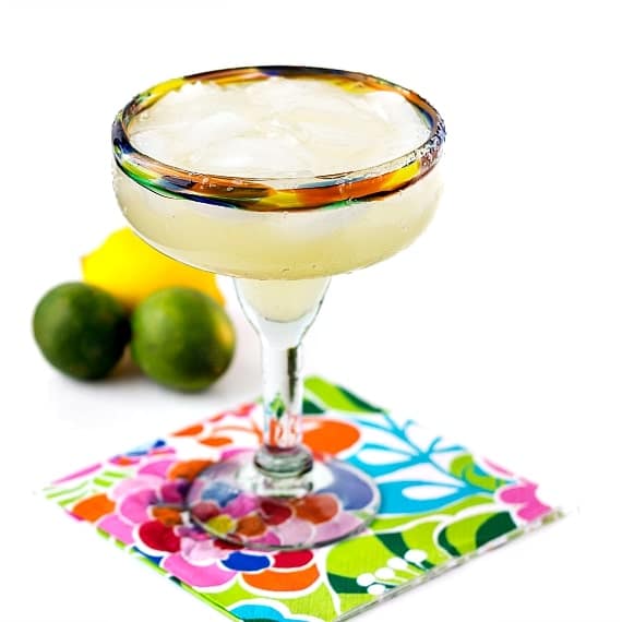 A top shelf margarita with a salted rim on a floral napkin with 2 limes and 1 lemon in the background.