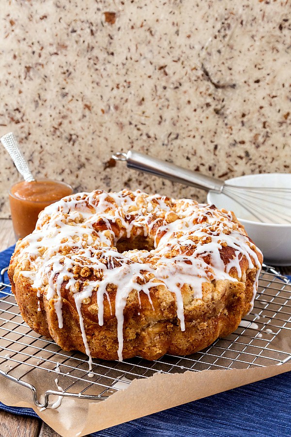 A large round loaf of apple butter coffee cake with white glaze on a cooling rack with apple butter and a bowl of glaze in the background.