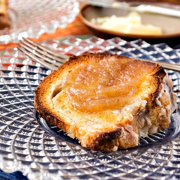 Close up of a slice of toasted apple butter coffee cake spread with even more apple butter on a glass plate with butter in the background.