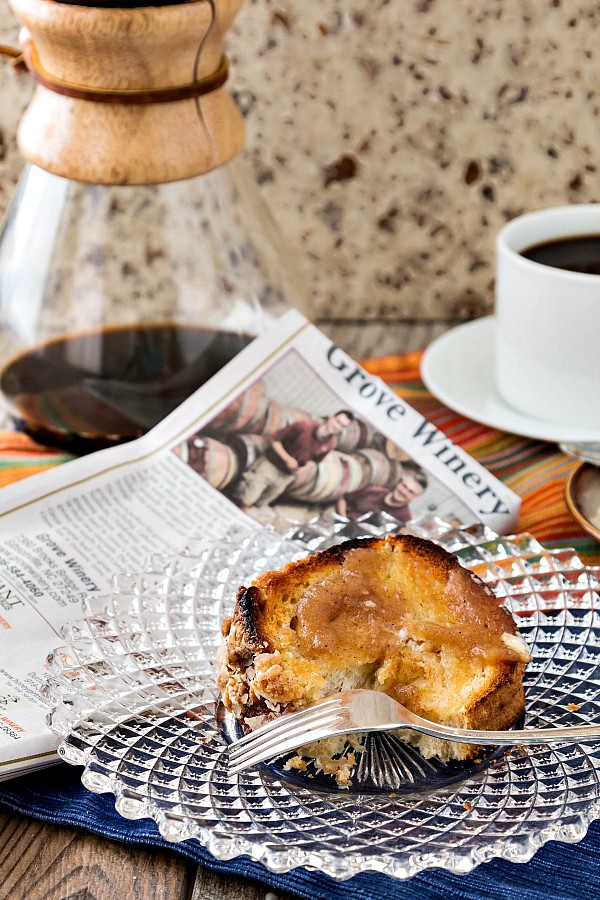 A slice of toasted yeast raised coffee cake on a glass plate along side a newspaper and a carafe of hot coffee.