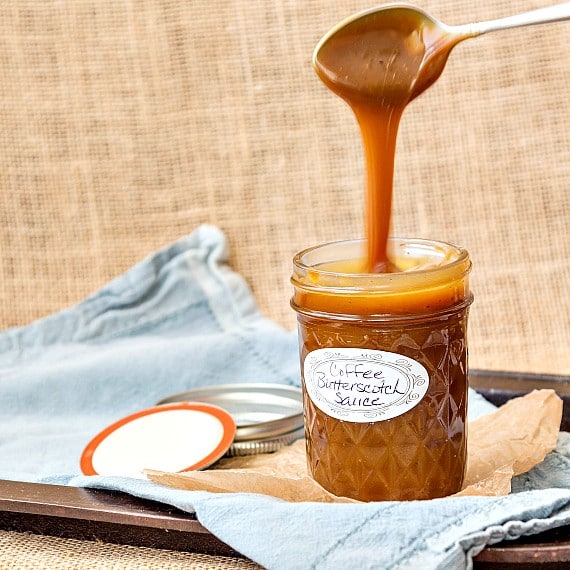 A jar of ice cream sauce with spoon lifted up and sauce drizzling back into the jar. Jar is a small mason jar and labeled "coffee butterscotch sauce". Ring top is next to it, on a linen faded blue napkin with a burlap background.