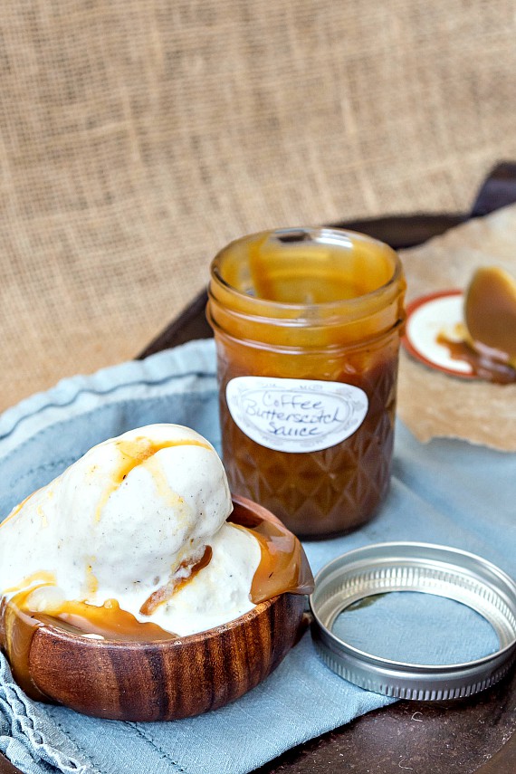 A small mason jar of ice cream sauce with a label reading "coffee butterscotch sauce". Next to a wooden bowl of ice cream, just starting to melt, with coffee butterscotch sauce melting into it. 