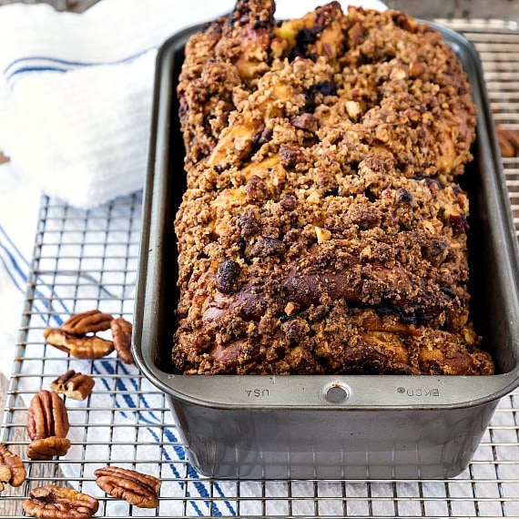 A loaf of sweet bread with streusel topping in a bread pan on a cooling rack with pecans.