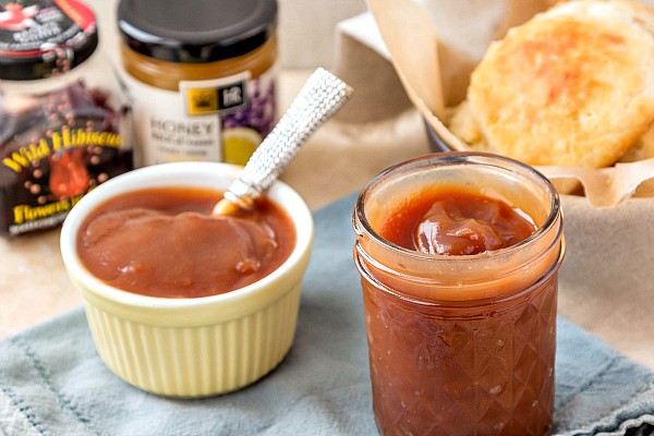 A jar and ramekin filled with a homemade honey apple butter recipe with biscuits in the background.