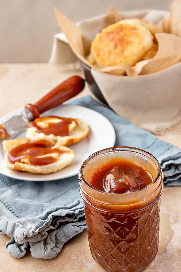 Jar of apple butter with a basket of biscuits and a split biscuit spread with apple butter in the background.