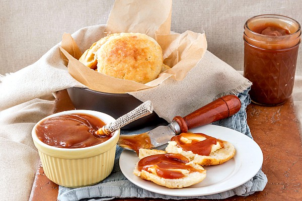 A ramekin of apple butter next to a plate with a split biscuit, slathered with apple butter. 