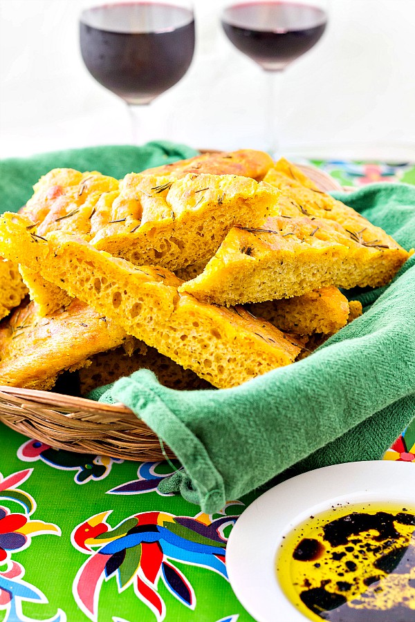 A basket of herbed pumpkin focaccia  cut into triangles with 2 glasses of wine in the background. A dish of dipping oil and vinegar is ready alongside the basket.