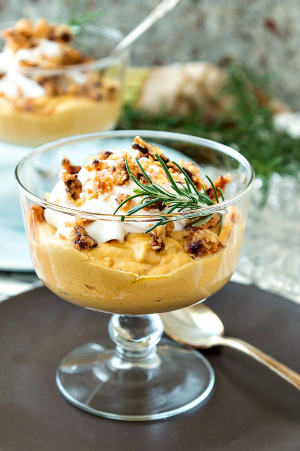 Close up a a footed glass bowl of pumpkin mousse on a brown plate with a spoon.