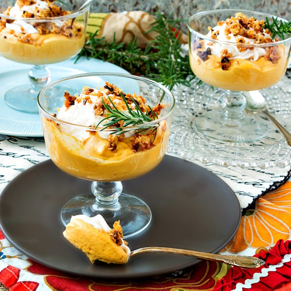 Individual footed clear bowls of caramel pumpkin mousse topped with whipped cream, streusel, and sprigs of rosemary.