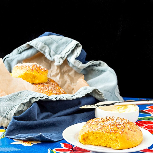 A pumpkin dinner roll on a plate with a basket of rolls in the background. One bite is missing from soft fresh roll.