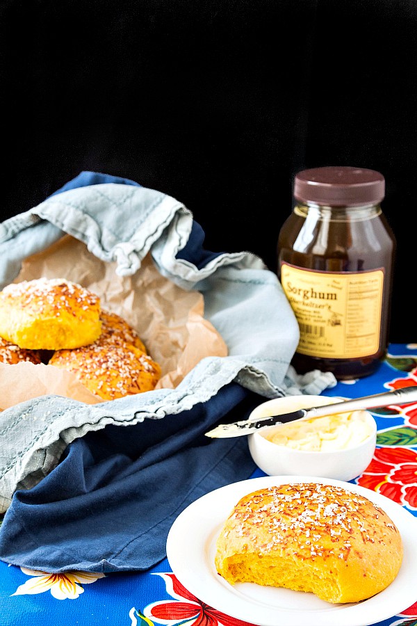 Soft pumpkin dinner rolls in a basket with a bread plate and a small dish of butter and a jar of sorghum syrup.