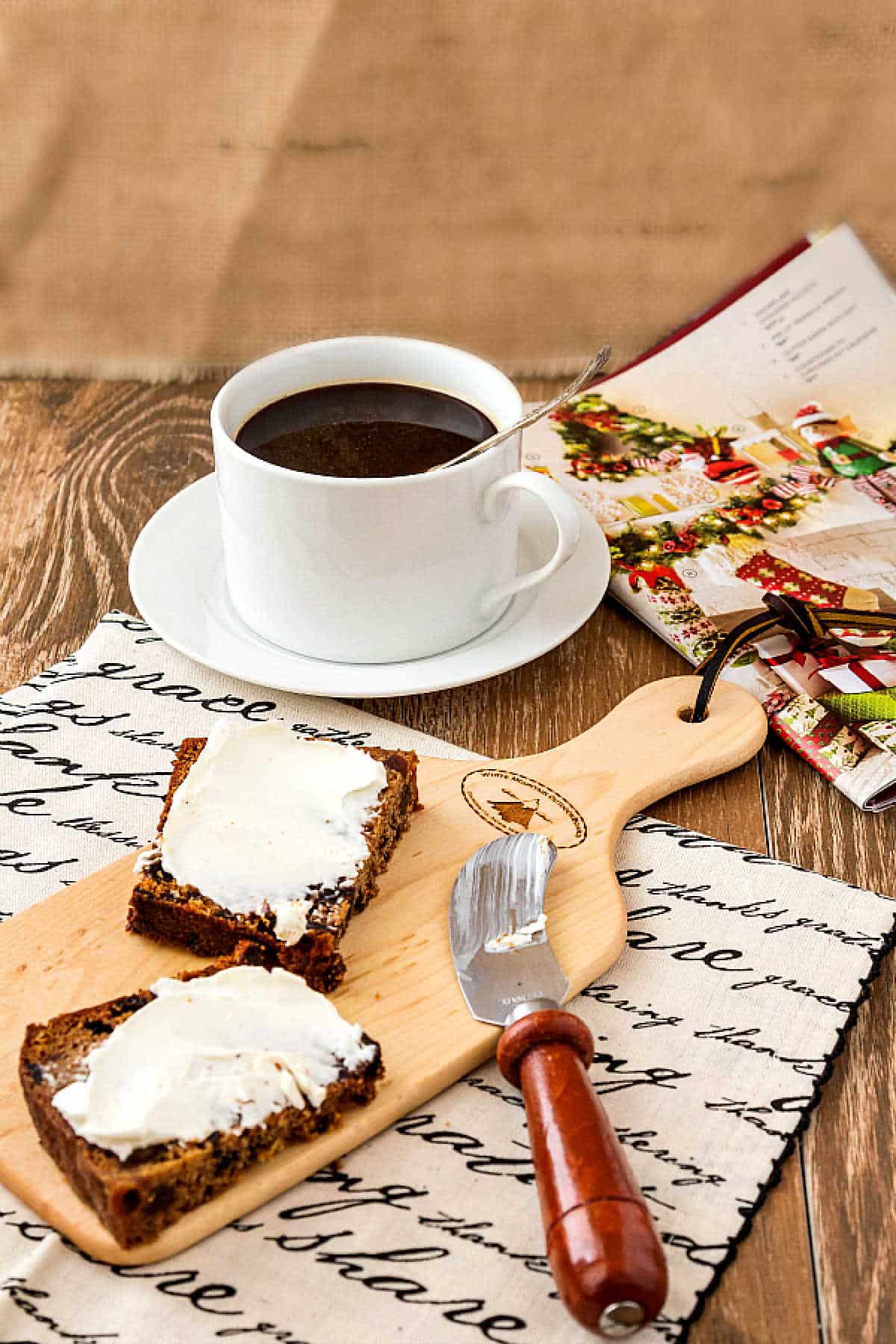 Sliced fruitcake spread with cream cheese on a small cutting board with a cup of coffee in the background.