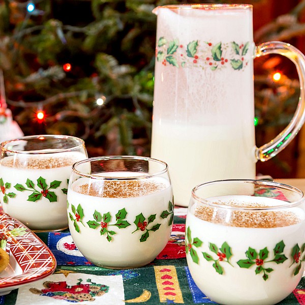 3 holiday glasses of eggnog, each sprinkled with nutmeg, with a pitcher of eggnog, all in front of a Christmas tree.