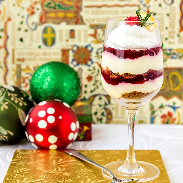 A wine glass layered with orange cheesecake mix, cranberry orange jam, and gingersnap crumbles. Christmas decorations in the background.