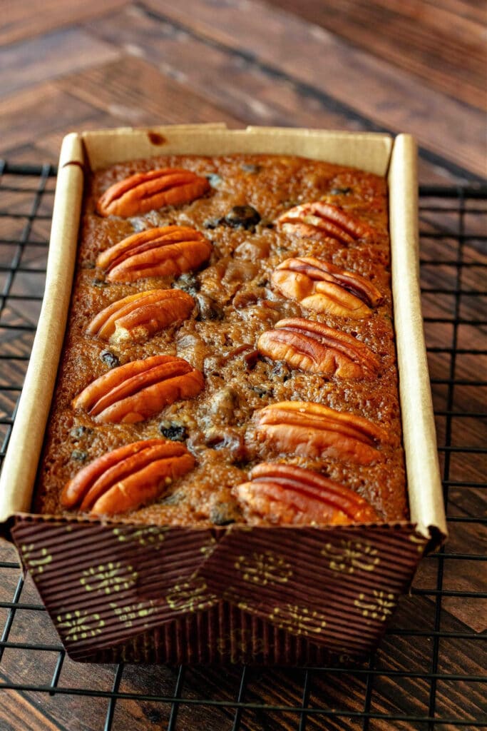 A whole fruitcake decorated with pecans on top in a paper loaf pan on a cooling rack.