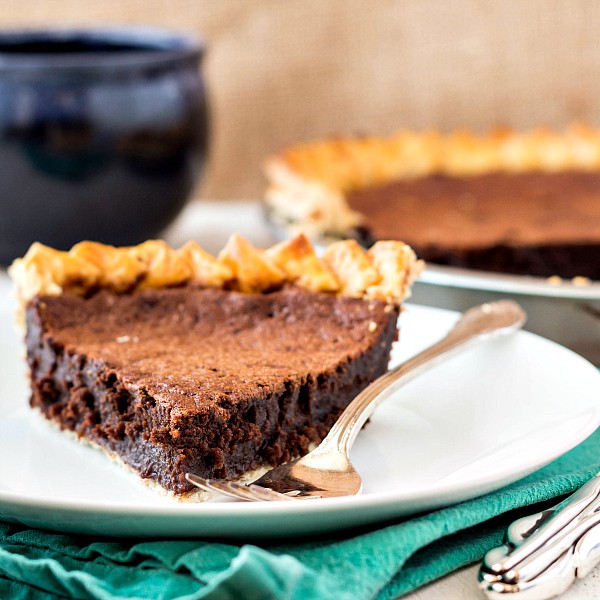 a slice of an old fashioned pie recipe: chocolate chess pie on a white plate with a blue coffee mug in the background