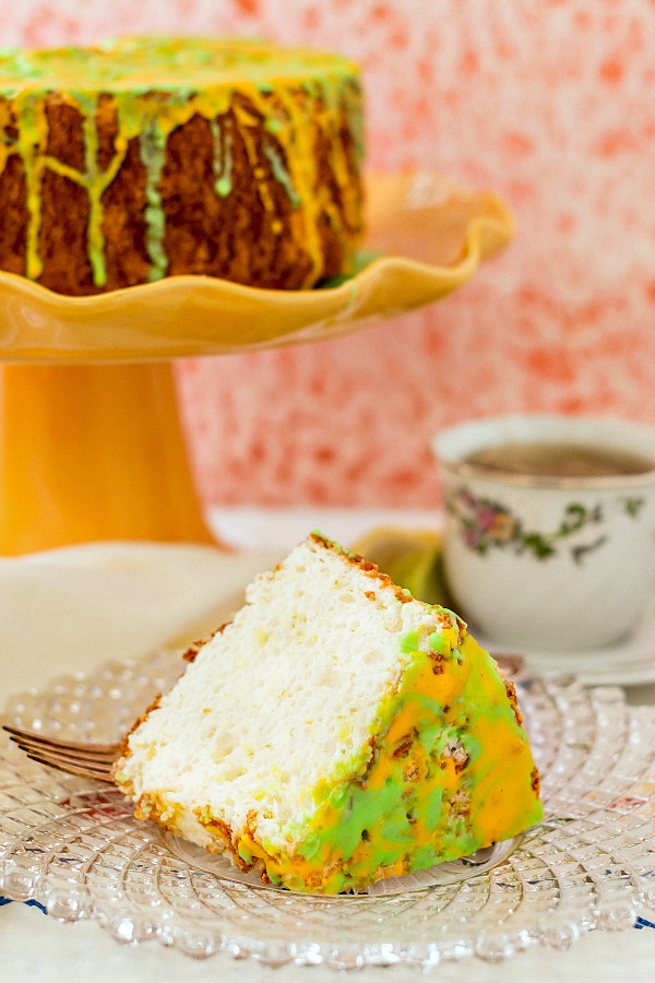 An angel food cake on a yellow cake stand and a slice of the cake on a plate with a fork and a cup of tea. 