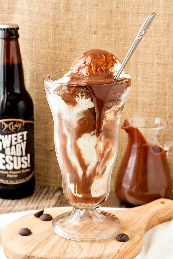 A tall sundae glass filled with ice cream and chocolate peanut butter sauce with a long metal spoon and a bottle of Sweet Baby Jesus in the background.