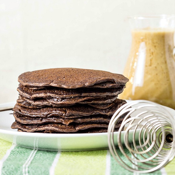 A plate of stacked chocolate yeast raised pancakes on a white plate with a whisk and a pitcher of coffee maple syrup in the background.