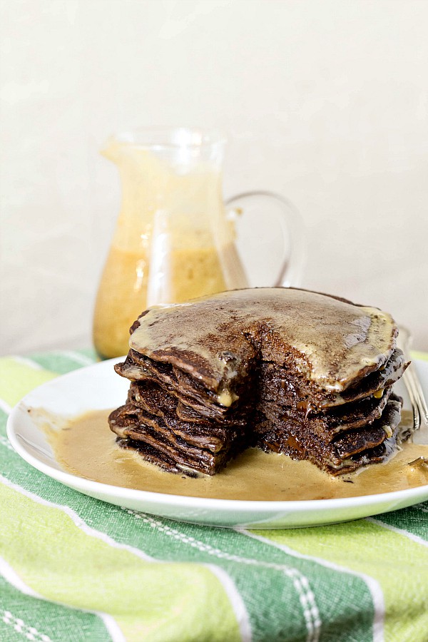 A tall stack of chocolate pancakes with chocolate chips on a white plate with syrup poured over them and a pitcher of coffee maple syrup in the background.