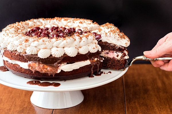 A whole fudgy black forest cake on a white footed stand. A hand with a serving knife is removing one serving.