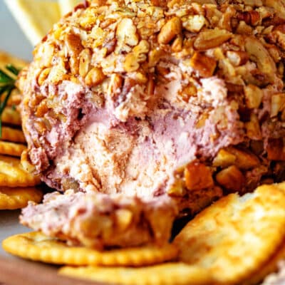 A cheeseball on a plate surrounded by crackers. Some of the cheeseball has been cut and is spread on one of the crackers.