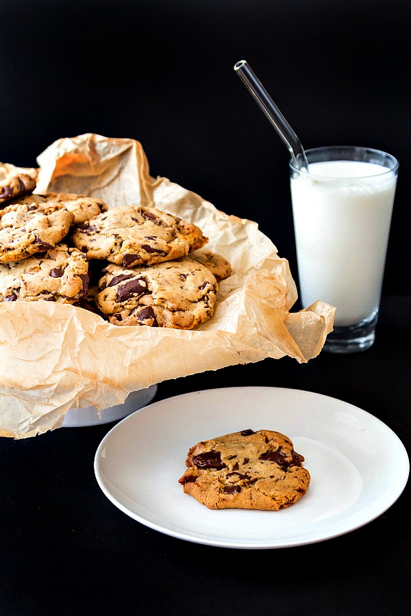 A plate of chocolate chunk cookies and a glass of milk with a glass straw in it. One cookie is on a single plate ready for serving.