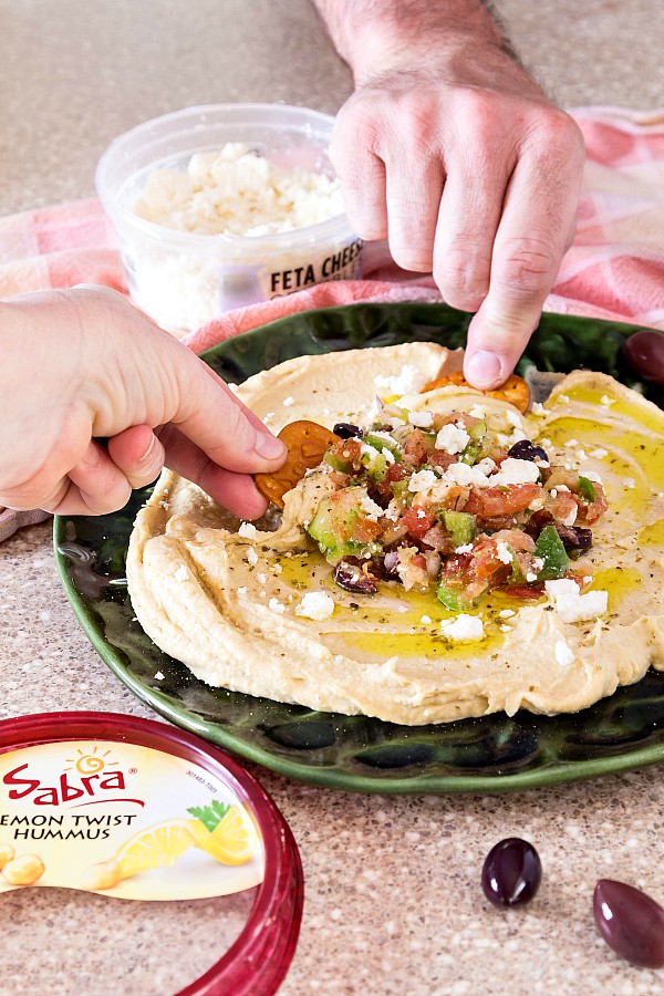 A plate with hummus spread on it with hummus toppings of chopped Greek salad, olive oil, and feta and crackers being dipped in for a bite.