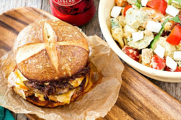 A burger on a pretzel bun next to a summer time salad.