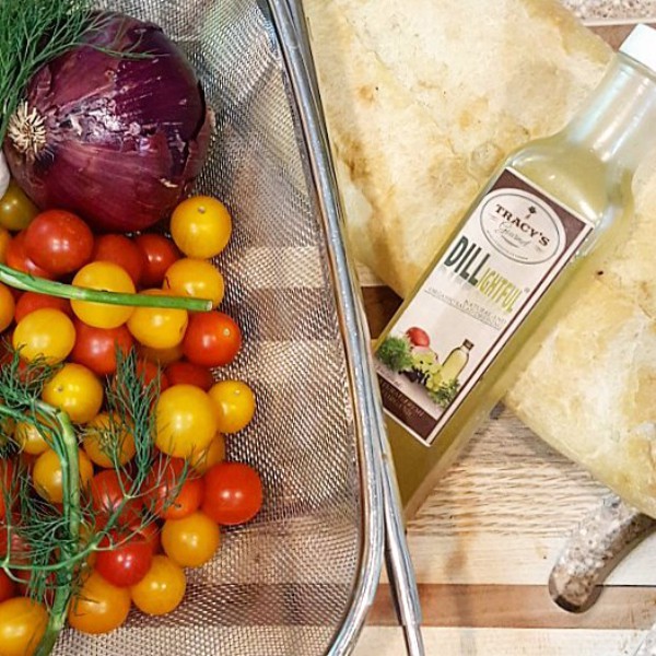 A colander of cherry tomatoes a whole red onion, and fresh dill next to a bottle of Tracy's Dill-lightful dressing.