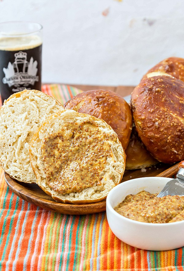 A wooden platter of pretzel buns. One is split open and slathered with whole grain mustard. A glass of dark beer is in the background.